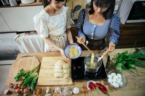 Free High angle crop content Asian females in stylish wear boiling homemade noodles in saucepan while cooking traditional Asian food together in contemporary kitchen Stock Photo