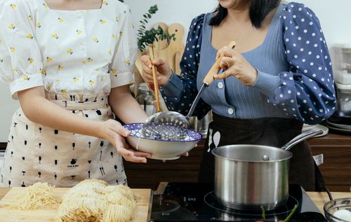 Free Crop anonymous housewives in stylish wear putting homemade boiled noodles into bowl while cooking together in modern kitchen Stock Photo
