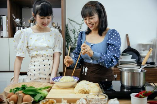 Free Smiling content Asian females wearing stylish clothes and aprons with chopsticks cooking yummy noodles on kitchen counter Stock Photo
