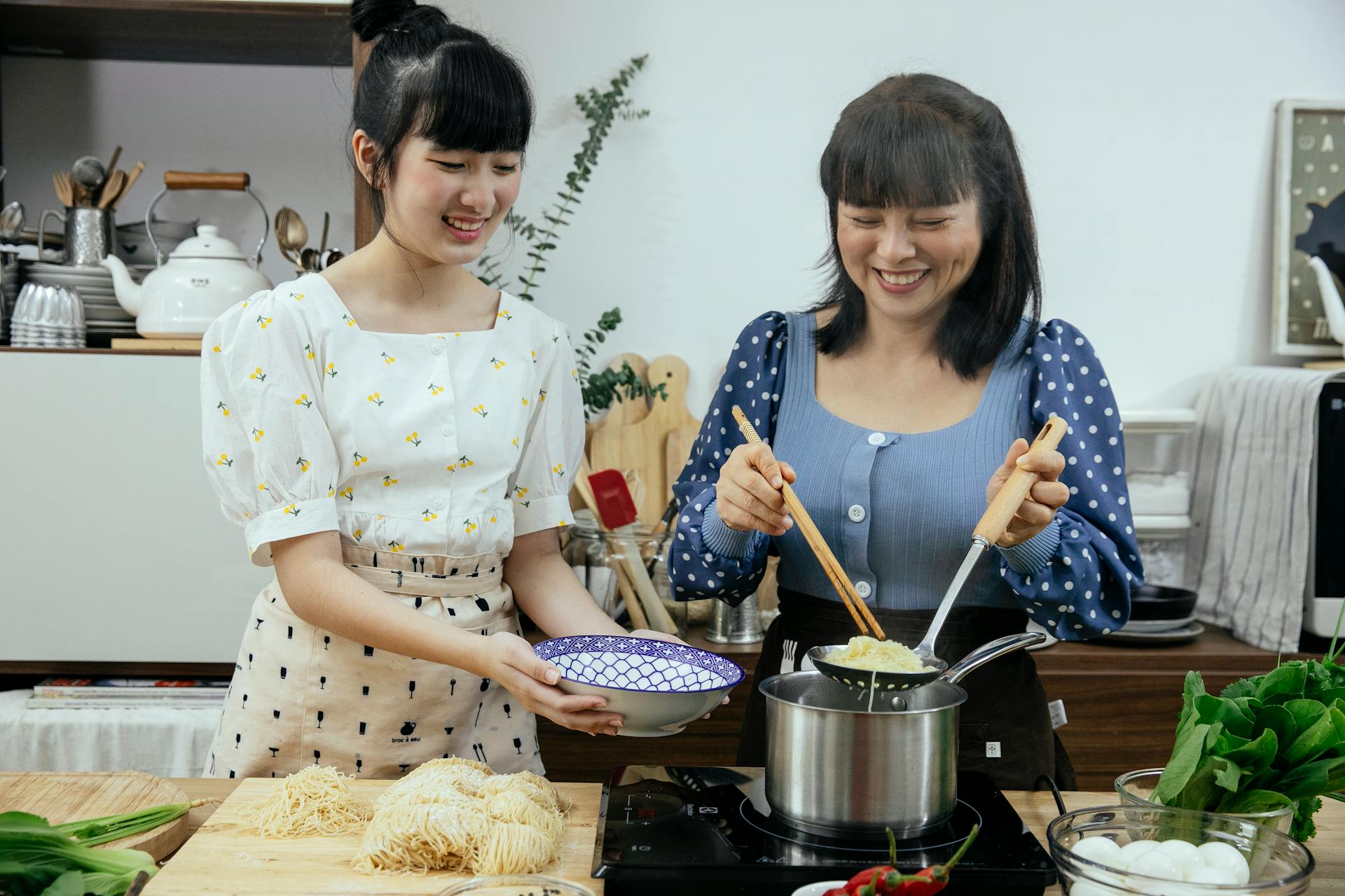 Happy Asian females in stylish clothes and aprons putting tasty boiled noodles into bowl while cooking traditional Asian dishes together in modern kitchen