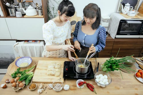 High angle glad Asian females in stylish aprons with chopsticks boiling homemade noodles in saucepan on tabletop stove while cooking Asian dish together in contemporary kitchen