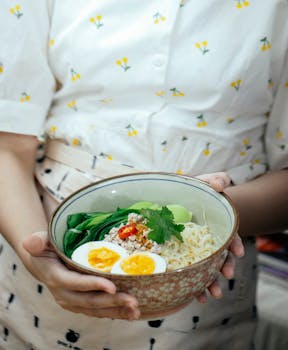 Crop anonymous female in apron showing bowl with delicious Asian ramen soup with noodles boiled egg and pak choi cabbage