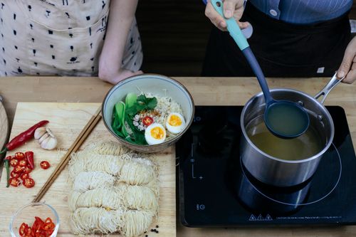 Free High angle crop anonymous females adding hot fresh vegetable broth to traditional Asian noodles soup in bowl while cooking together in modern kitchen Stock Photo