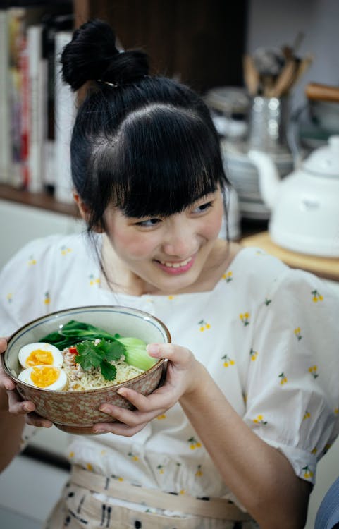 Happy smiling Asian female in stylish wear demonstrating freshly cooked Asian noodles soup in bowl and looking away contentedly