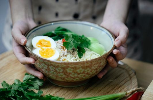 Crop anonymous female touching bowl with delicious freshly cooked Asian noodles soup with boiled egg and parsley placed on round wooden board