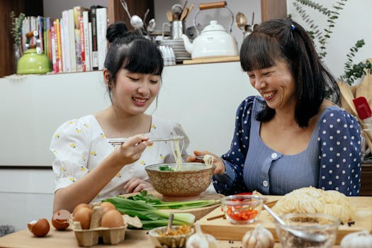 Two women smiling and enjoying a noodle meal together in a kitchen setting.
