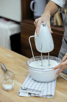 Crop anonymous female chef beating eggs and preparing fluffy whipped cream with mixer while baking in kitchen