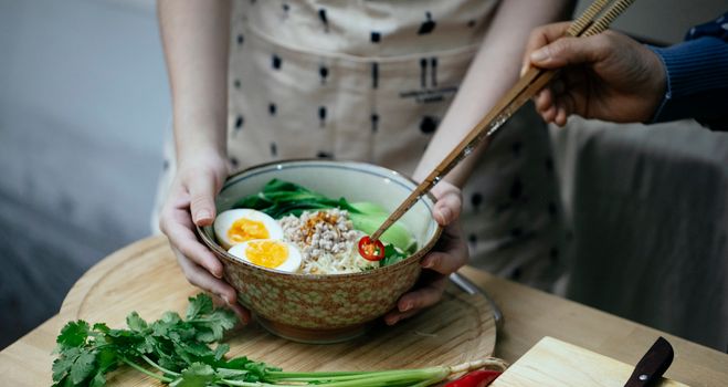 Crop anonymous females garnishing with cut chili pepper appetizing traditional Asian noodles soup with boiled egg and pak choi cabbage while cooking together in kitchen
