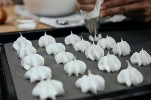Crop unrecognizable chef with piping bag with star tip forming vanilla meringue cookies on baking pan in kitchen