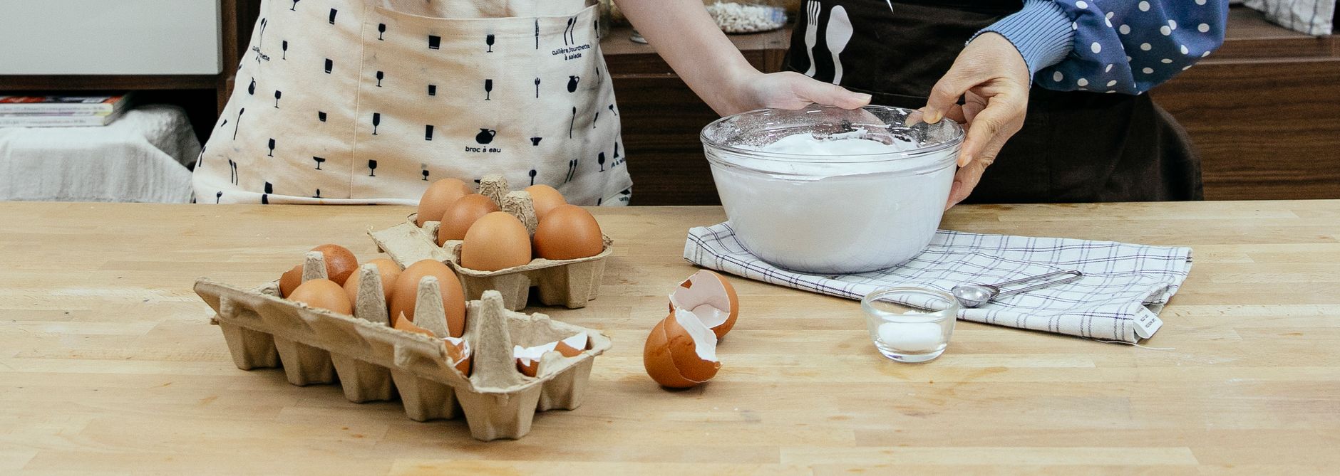 Crop unrecognizable females wearing aprons touching bowl with fresh whipped cream placed on table near eggs in modern kitchen