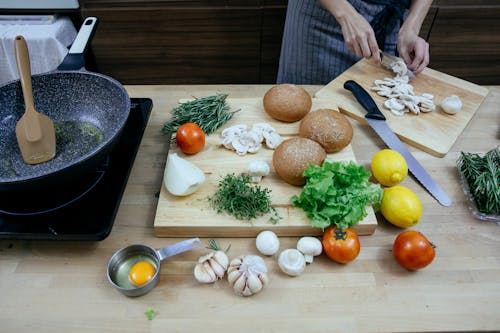 Crop anonymous female chef wearing apron cutting fresh mushrooms while standing at table with scattered ripe vegetables including onions tomatoes and potatoes in modern kitchen