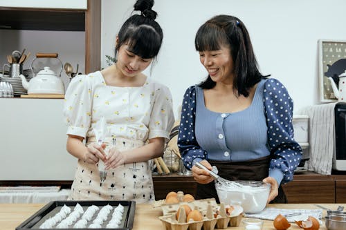 Happy middle aged Asian female in casual clothes with teenage daughter smiling while preparing tasty meringue cookies in kitchen