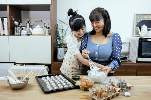 Happy Asian mother and daughter cuddling while preparing meringues in kitchen