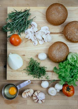 Top view of fresh mushrooms with tomato and onion arranged on wooden cutting board with bread buns and assorted herbs placed near garlic and egg yolk during dinner preparation