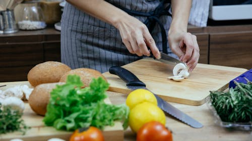 Free Crop anonymous female in apron cutting fresh mushrooms on wooden chopping board while preparing dinner with vegetable and herbs in kitchen Stock Photo