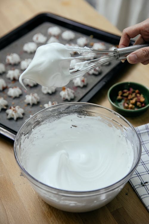 From above of anonymous female cook with whipped egg whites in bowl placed on table with meringue cookies on tray on blurred background