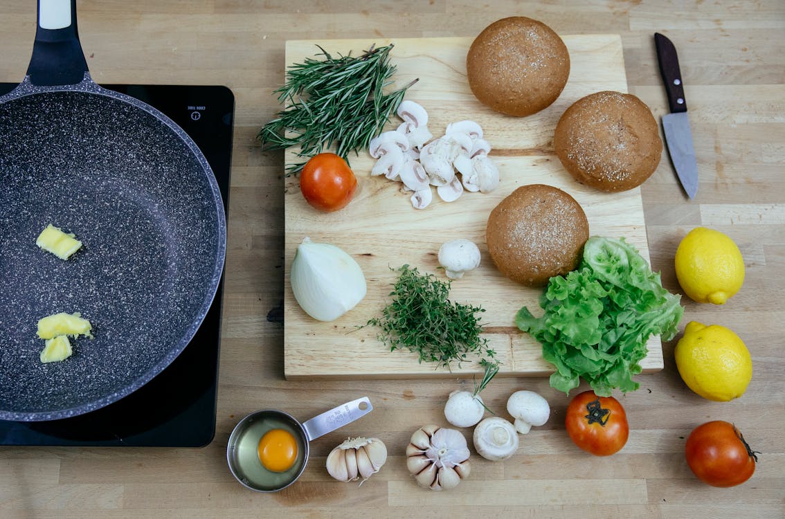Free Top view of pan with butter on stove placed near wooden board with buns greens and fresh vegetables in kitchen Stock Photo