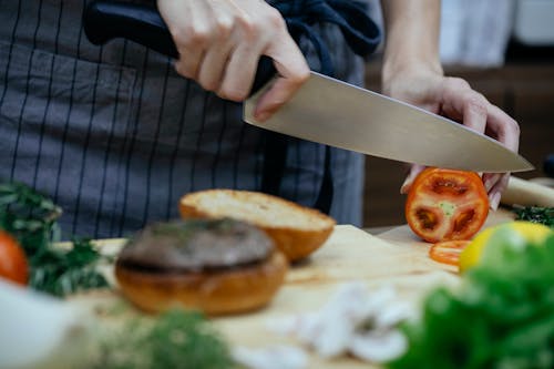 Free Unrecognizable female cook cutting fresh tomato with knife while standing at table with toasted buns on wooden cutting board while cooking against blurred background Stock Photo