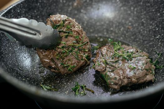 Juicy cutlets topped with aromatic rosemary frying in hot pan with metal tongs during cooking process in kitchen while preparing for lunch