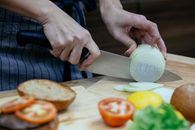 Unrecognizable female cook slicing onion on cutting board at table with blurred tomatoes and toasted buns in kitchen during cooking process