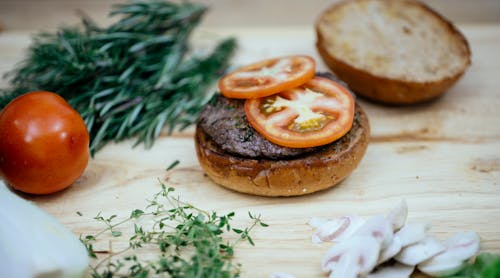 Free Toasted buns with cutlet and tomatoes placed on wooden table with greens and mushroom in kitchen while preparing tasty burger Stock Photo