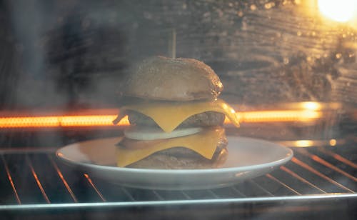 Through glass of tasty burger with cutlet and cheese served on white plate in hot oven on rack while roasting