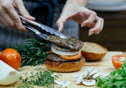 Unrecognizable female cook using tongs to put juicy meat on bun with onion and slice of tomato while making burger against blurred background