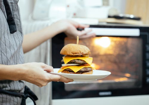Free Unrecognizable person with plate of burger in hand opening door of oven to roast food while cooking in kitchen on blurred background Stock Photo