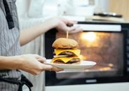 Unrecognizable person with plate of burger in hand opening door of oven to roast food while cooking in kitchen on blurred background