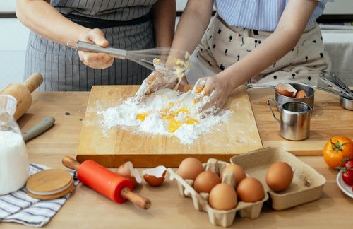 Free From above of unrecognizable female cooks in aprons cooking at table with ingredients and wooden board while mixing ingredients for dough in kitchen Stock Photo