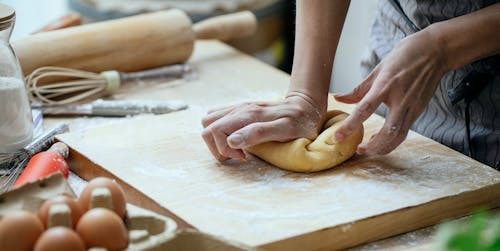 Free Unrecognizable female cook pressing piece of dough on wooden board while cooking at table with whisk and rolling pin against blurred background Stock Photo