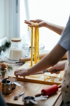 Unrecognizable cooks with long raw homemade pasta standing near table with cutting board and kitchenware while cutting dough in kitchen on blurred background