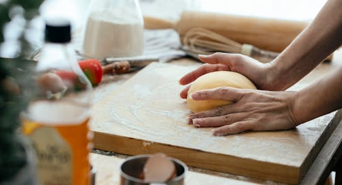 Anonymous female cook with ball of raw dough on wooden board standing at table while kitchenware while cooking in kitchen against blurred background