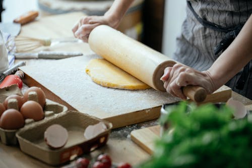 Unrecognizable female cook flattening dough with rolling pin while standing at table with wooden board against blurred background in kitchen