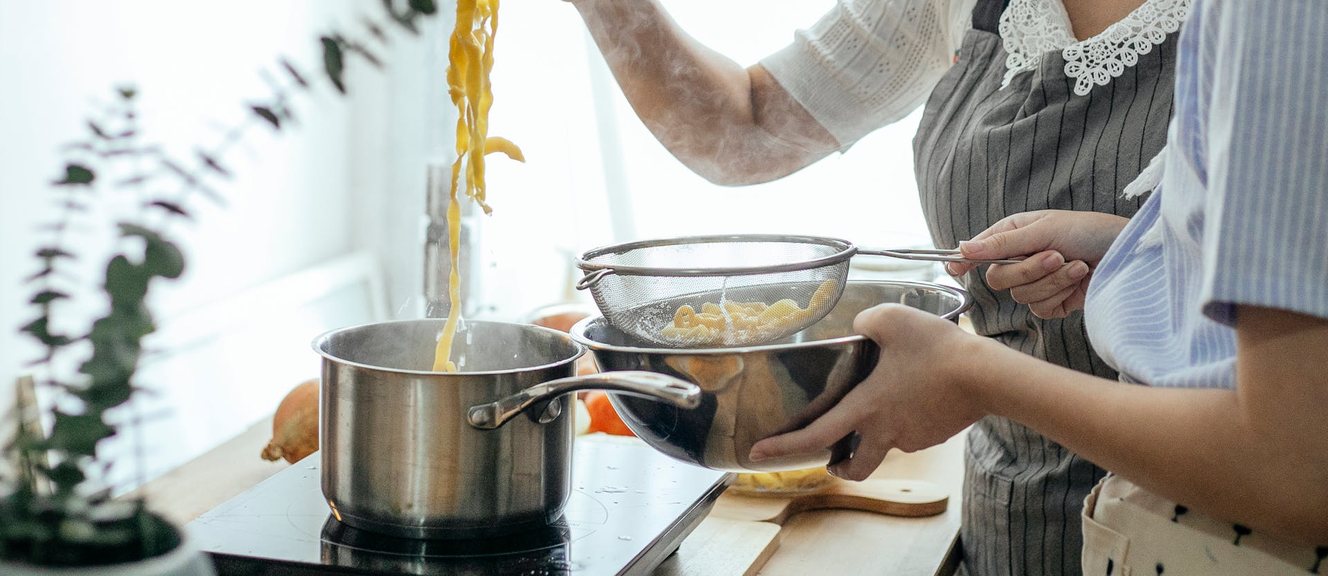 Unrecognizable female cooks putting pasta from pot into sieve while cooking lunch at table with stove in kitchen on blurred background