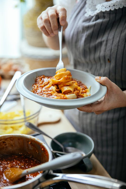 Free Unrecognizable female cook with plate of pasta wrapped around fork standing at table with pan of tasty chili against blurred background Stock Photo