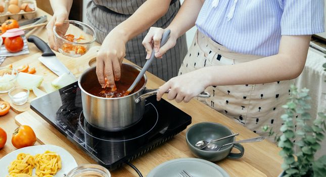 From above of anonymous female cooks adding chopped tomatoes into saucepan with boiling chili while standing at table with stove