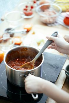 From above of unrecognizable person stirring tasty chili in saucepan with spoon while cooking on stove at table in kitchen on blurred background