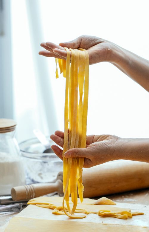 Free Unrecognizable person with long uncooked pasta standing at table with rolling pin while cooking tagliatelle with dough in kitchen against blurred background Stock Photo