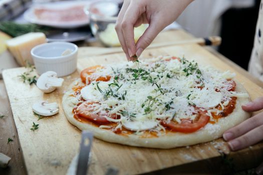 Unrecognizable female cook putting fresh greens on uncooked pizza with sliced tomatoes and mushrooms topped with grated cheese while cooking against blurred background