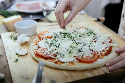 Free Unrecognizable female cook putting fresh greens on uncooked pizza with sliced tomatoes and mushrooms topped with grated cheese while cooking against blurred background Stock Photo