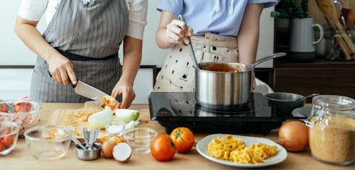 Unrecognizable woman cutting fresh tomato on cutting board while standing at table with ingredients and stove near female cook stirring sauce in pan