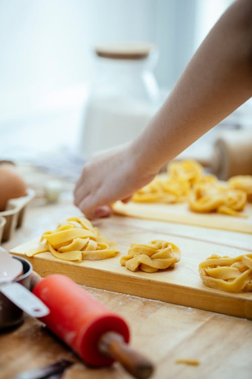 Unrecognizable cook with tagliatelle nests on wooden cutting board standing at table while preparing dough for pasta against blurred background