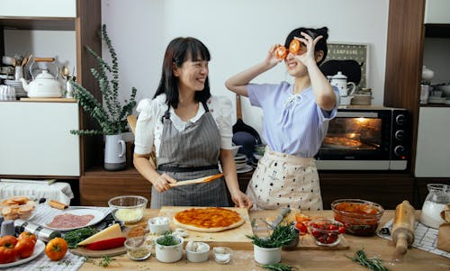 Free Smiling Asian woman spreading tomato sauce on pizza dough while looking at funny female covering eyes with tomato slices in kitchen Stock Photo
