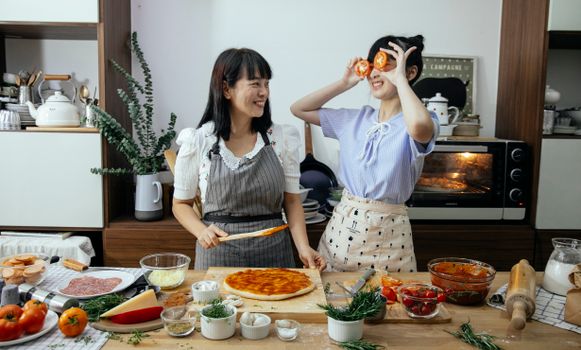一位微笑的亚洲女子正在往披萨面团上涂番茄酱，同时看着厨房里那位用番茄片遮住眼睛的有趣女子