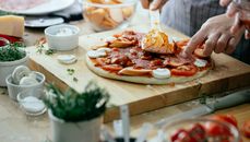 Unrecognizable chef spreading sauce on pizza with tomatoes mushrooms and salami placed on wooden cutting board on table with ingredients