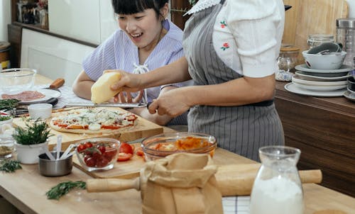 Happy Asian woman at table with unrecognizable woman rubbing cheese on uncooked pizza in kitchen with various ingredients during cooking process