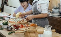 Happy Asian woman at table with unrecognizable woman rubbing cheese on uncooked pizza in kitchen with various ingredients during cooking process