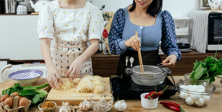 Unrecognizable female cooks standing at table with various ingredients and raw pasta on cutting board while cooking noodles on stove in pan