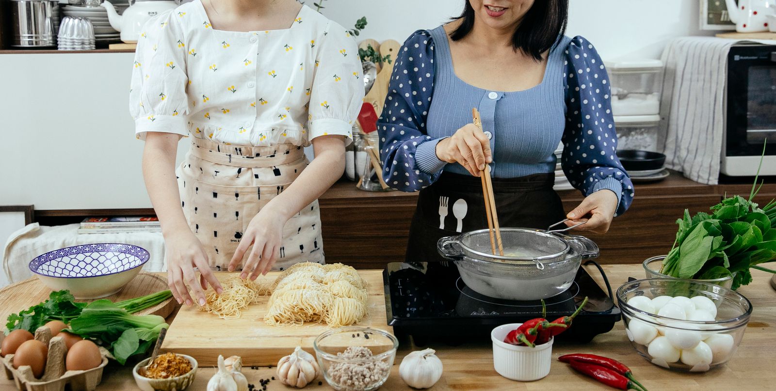Unrecognizable female cooks standing at table with various ingredients and raw pasta on cutting board while cooking noodles on stove in pan